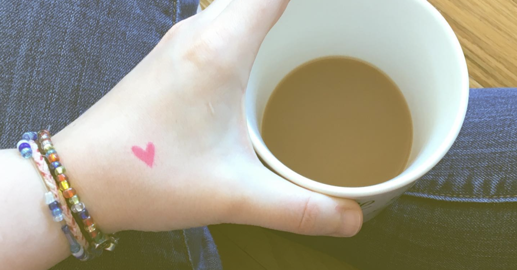 An aerial image of white person's hand holding a white mug of tea. There is a small pink heart between her thumb and forefinger. She is wearing friendship bracelets. You can see crossed legs wearing jeans in the background, and she is sitting on a wooden floor. 